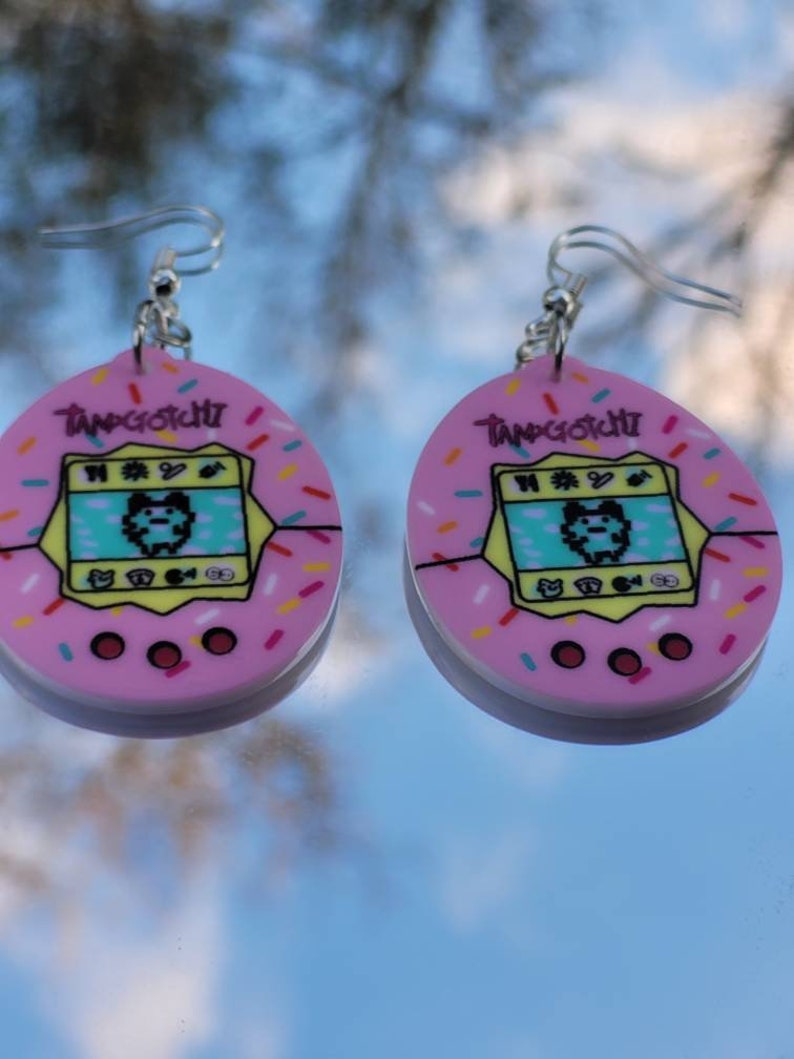 Może przedstawiać: Para r&oacute;żowych kolczyk&oacute;w Tamagotchi. Każdy kolczyk ma r&oacute;żowy owalny kształt z ż&oacute;łtą obw&oacute;dką i kolorową posypką. Ekran Tamagotchi wyświetla spikselizowanego zwierzaka. Srebrne haczyki do uszu dopełniają design.
