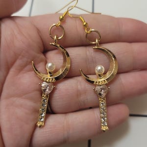 May include: Gold crescent moon earrings with a pearl and a pink crystal heart on a stick with rhinestones.