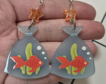 May include: A pair of novelty earrings featuring a red goldfish in a blue plastic bag design. The earrings have orange star-shaped accents and silver-colored hooks. The fish have yellow seaweed and white bubbles.