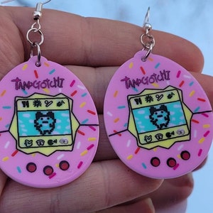 Może przedstawiać: R&oacute;żowe kolczyki Tamagotchi z posypką. Każdy kolczyk ma retro cyfrowy wz&oacute;r zwierzaka z napisem "Tamagotchi". Kolczyki mają srebrne zapięcia na haczyk.