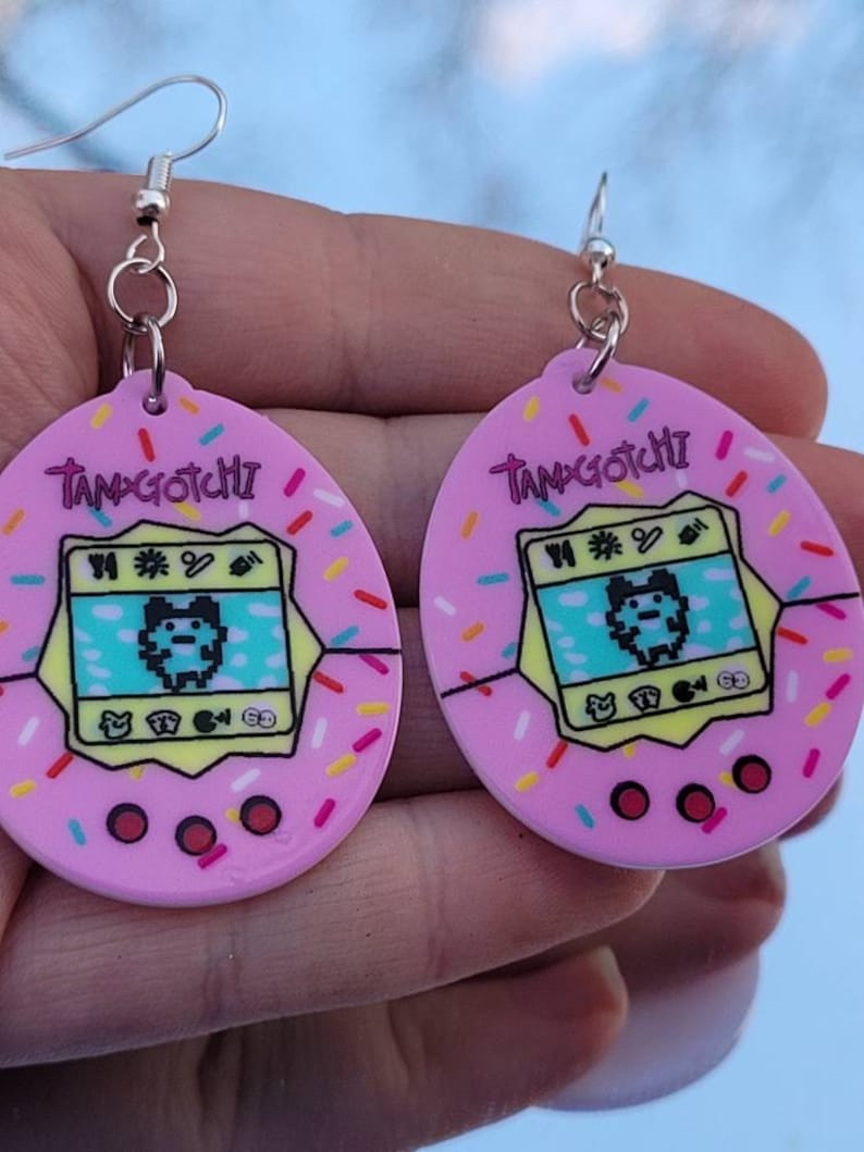 Może przedstawiać: R&oacute;żowe kolczyki Tamagotchi z posypką. Każdy kolczyk ma retro cyfrowy wz&oacute;r zwierzaka z napisem "Tamagotchi". Kolczyki mają srebrne zapięcia na haczyk.