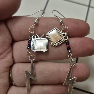 Puede incluir: Pendientes plateados con un diseño de televisión retro y un dije de rayo. Los pendientes tienen una cadena y detalles de cuentas con cuentas rojas, azules y negras. Los pendientes cuelgan de un gancho plateado.