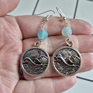 May include: Silver-toned dangle earrings. Each earring features a round pendant with a wave design and a blue bead. The pendants have an iridescent, pearlescent finish. The earrings are held by a hand.