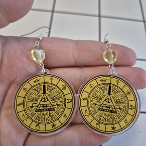 May include: A pair of dangle earrings featuring a yellow circular design with a black illustration of a triangle eye and other symbols. The earrings have silver hooks and a heart-shaped bead.