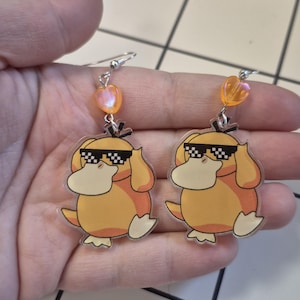 May include: A pair of dangle earrings featuring a cartoon duck wearing sunglasses. The earrings are yellow and orange with silver hooks and heart-shaped orange beads. The duck has a white belly and is wearing black and white checkered sunglasses.