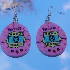 Może przedstawiać: Para r&oacute;żowych kolczyk&oacute;w Tamagotchi. Każdy kolczyk ma r&oacute;żowy owalny kształt z wzorem ekranu Tamagotchi, słowem "Tamagotchi" na g&oacute;rze i kolorowymi posypkami na krawędzi. Kolczyki mają srebrne zapięcia na haczyk.