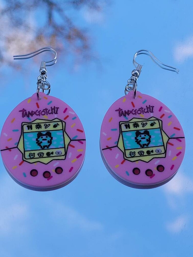 Może przedstawiać: Para r&oacute;żowych kolczyk&oacute;w Tamagotchi. Każdy kolczyk ma r&oacute;żowy owalny kształt z wzorem ekranu Tamagotchi, słowem "Tamagotchi" na g&oacute;rze i kolorowymi posypkami na krawędzi. Kolczyki mają srebrne zapięcia na haczyk.