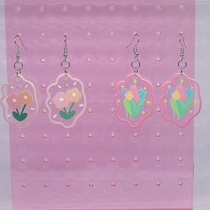 May include: Four pairs of pink resin earrings with flower and floral designs. The earrings are displayed on a pink pegboard.