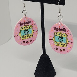 Może przedstawiać: Para r&oacute;żowych kolczyk&oacute;w z motywem Tamagotchi. Każdy kolczyk ma r&oacute;żowy owalny kształt z kolorowym wzorem postaci Tamagotchi i napisem "Tamagotchi". Kolczyki mają srebrne haczyki.