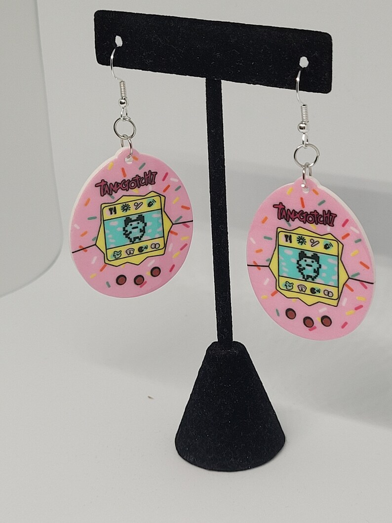 Może przedstawiać: Para r&oacute;żowych kolczyk&oacute;w z motywem Tamagotchi. Każdy kolczyk ma r&oacute;żowy owalny kształt z kolorowym wzorem postaci Tamagotchi i napisem "Tamagotchi". Kolczyki mają srebrne haczyki.
