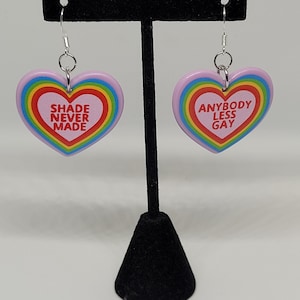 May include: A pair of rainbow heart-shaped earrings with the text "Shade Never Made" on one earring and "Anybody Less Gay" on the other earring.