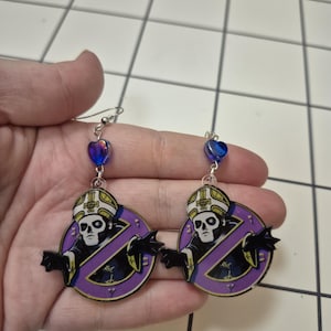 May include: A pair of dangle earrings featuring a skull-faced figure in a purple circle with a no-entry symbol. The earrings have silver hooks and a blue heart-shaped bead at the top.