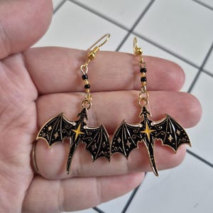 May include: A pair of gold-colored dragon earrings. Each earring features a black dragon with outstretched wings, detailed with gold accents and a yellow starburst. The earrings hang from gold-colored hooks with black beads.