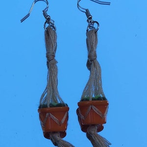 Peut inclure: Une paire de boucles d'oreilles pendantes avec de petits pots orange contenant des plantes vertes suspendus à des cordons en macramé beige.