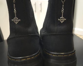 May include: A pair of black leather ankle boots with a chunky sole. Each boot has a silver charm with a cross design hanging from a ring at the top. The boots have yellow stitching around the sole.