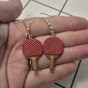 Puede incluir: Un par de pendientes de raqueta de ping-pong rojos con ganchos plateados. Las raquetas tienen una superficie texturizada y mangos marrones. Los pendientes están sostenidos en una mano.