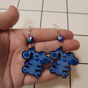 May include: A pair of dangle earrings featuring a blue cartoon dog with iridescent purple heart charms.