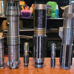 May include: A collection of eight 3D printed lightsaber hilts, each with a unique design and name. The hilts are made of a silver-colored plastic and have various details, including buttons, switches, and grooves. The hilts are arranged in a row on a wooden surface. The names on the hilts are "Halley", "Priscilla", "Cathryn", "Anakin", "Lando", "Ezra", and "Ahsoka".