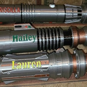 May include: Three silver lightsaber hilts with personalized names: Priscilla, Hailey, and Lauren. The hilts have a copper-colored accent and a black band.