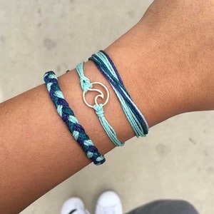 May include: A set of three bracelets with a wave charm. The bracelets are made of blue and green string and are tied together with a silver charm.