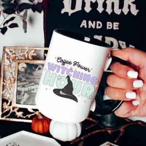 Könnte beinhalten: Eine weiße Keramiktasse mit schwarzem Henkel. Die Tasse zeigt ein Motiv eines Hexenhutes mit dem Text "Coffee Power Witching Hour" in bunten Buchstaben.