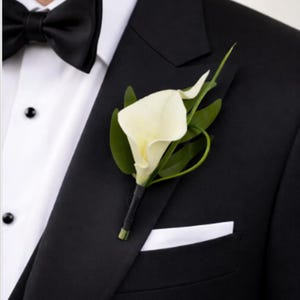 Puede incluir: Un boutonniere de cala blanca con hojas verdes prendido en la solapa de una chaqueta de traje negra. El traje se lleva con una camisa blanca, una pajarita negra y un pañuelo de bolsillo blanco en el bolsillo del pecho.