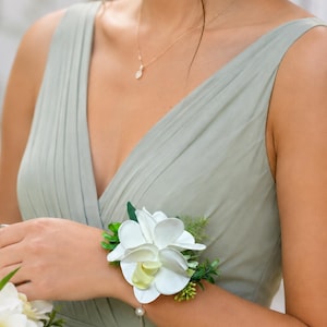 May include: A wrist corsage featuring white orchid blossoms with pale yellow centers, accented by small green leaves and sprigs, attached to a delicate bracelet with a single white pearl bead, worn with a sage green sleeveless dress.