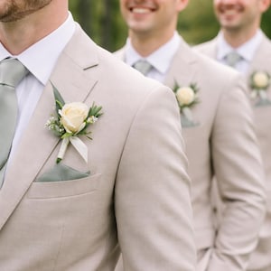 Op de afbeelding: Drie mannen in lichtbruine pakken met witte overhemden en lichtgroene stropdassen. Elk pak heeft een crème rozenknop en een bijpassende pochet. De mannen glimlachen, mogelijk bij een bruiloft of formeel evenement.