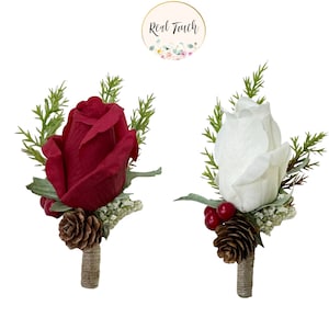 May include: Two artificial flower boutonnieres with a red rose and a white rose. Each boutonniere has pine sprigs, a pine cone, and red berries.