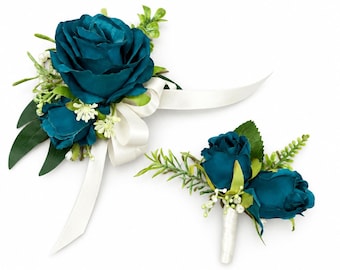 May include: A teal rose corsage and a matching boutonniere featuring deep teal fabric roses, small white filler flowers, and green leaves. The corsage includes a white satin ribbon, while the boutonniere has a white wrapped stem.