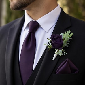 May include: A black suit jacket and vest worn over a crisp white dress shirt with a solid dark purple necktie. A matching dark purple pocket square is tucked into the jacket pocket, and a boutonniere features a dark purple rose with greenery.
