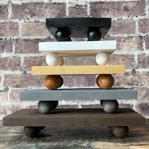 May include: A stack of five wooden risers in varying colors: black, white, yellow, gray, and brown. Each riser is supported by rounded wooden feet. The risers are arranged in a tiered fashion, creating a decorative display.