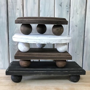 May include: A tiered wooden display stand with four levels. The levels are rectangular and round, painted in shades of brown, white, and black. Each level is supported by wooden ball feet, creating a rustic, farmhouse aesthetic.