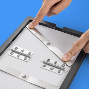 May include: A black frame with two white panels connected by silver hinges. The panels are held together by two silver metal bars. The frame is being held by two hands.