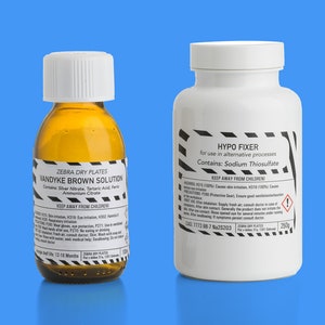 Può includere: Due bottiglie su sfondo blu. Una è in vetro ambrato con tappo bianco, etichettata "Vandyke Brown Solution". L'altra è una bottiglia di plastica bianca con tappo bianco, etichettata "Hypo Fixer". Entrambe le bottiglie hanno etichette di avvertimento.