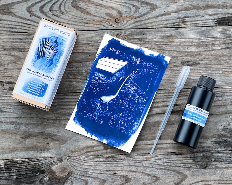 Op de afbeelding: Een blauwe cyanotype-afdruk van een houten constructie in een veld, een bruine doos met het label "The New Cyanotype", een zwarte fles cyanotype-oplossing en een doorzichtige plastic druppelaar.