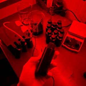 Op de afbeelding: Een rood verlichte laboratoriumwerkplek met verschillende glazen bekers en flessen. Een hand houdt een donkere glazen fles vast met een zwarte dop. Een witte verwarmings plaat met een zwarte knop staat op de tafel.