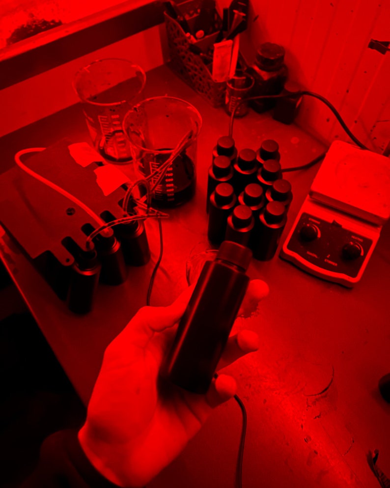 Op de afbeelding: Een rood verlichte laboratoriumwerkplek met verschillende glazen bekers en flessen. Een hand houdt een donkere glazen fles vast met een zwarte dop. Een witte verwarmings plaat met een zwarte knop staat op de tafel.