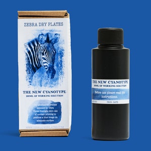 Op de afbeelding: Een zwarte fles met het label "The New Cyanotype 100ml of Working Solution" met instructies om te lezen voor gebruik. De fles staat naast een doos met het label "Zebra Dry Plates" met een blauwe en witte afbeelding van een zebra.