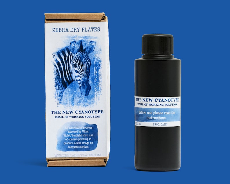 Op de afbeelding: Een zwarte fles met het label "The New Cyanotype 100ml of Working Solution" met instructies om te lezen voor gebruik. De fles staat naast een doos met het label "Zebra Dry Plates" met een blauwe en witte afbeelding van een zebra.