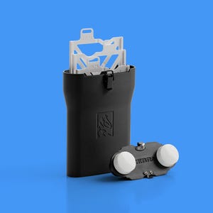 Peut inclure: Un étui de rangement rectangulaire noir avec un petit loquet, contenant des inserts de film en plastique blanc étiquetés FILM 1 et FILM 2. À côté de l'étui se trouve un composant noir avec deux boutons circulaires blancs et le texte ZEBRA.