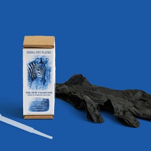 Op de afbeelding: Een bruine doos met de tekst "ZEBRA DRY PLATES THE NEW CYANOTYPE HOME OF WORKING SOLUTIONS" en een blauwe en witte afbeelding van een zebra. De doos staat op een blauwe ondergrond naast een paar zwarte nitril handschoenen en een doorzichtige plastic druppelaar.