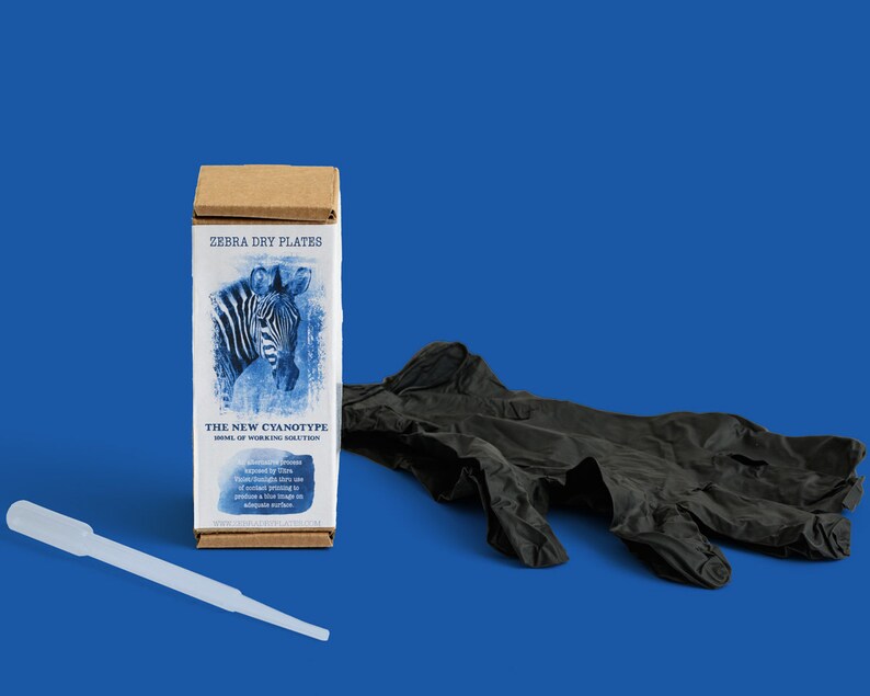 Op de afbeelding: Een bruine doos met de tekst "ZEBRA DRY PLATES THE NEW CYANOTYPE HOME OF WORKING SOLUTIONS" en een blauwe en witte afbeelding van een zebra. De doos staat op een blauwe ondergrond naast een paar zwarte nitril handschoenen en een doorzichtige plastic druppelaar.