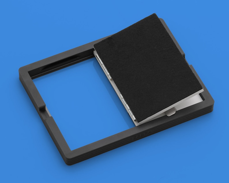 May include: A black frame with a black flap that opens to reveal a white interior. The frame is made of a textured material and has a raised edge. The flap is hinged and has a black fabric lining.