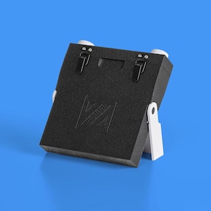 May include: A black, rectangular, foldable camera with a white stand. The camera has a textured surface and a logo on the front.