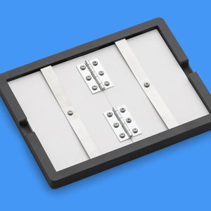 May include: A black framed folding board with two sets of silver hinges. The board is made of a light gray material and has a black rubber border.