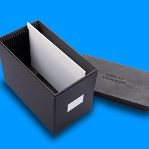 以下が含まれることがあります： 白い仕切りが付いた黒い収納ボックス。蓋が開いており、「ZEBRA 5x7 ARCHIVE BOX」と書かれています。