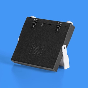 Może przedstawiać: Czarny, prostokątny futerał na aparat z białym stojakiem. Futerał ma logo z przodu i dwa zatrzaski na górze.