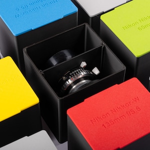 Peut inclure: Gros plan d'un objectif d'appareil photo à l'intérieur d'une boîte noire. L'objectif est entouré de boîtes colorées avec des étiquettes qui indiquent "Nikon Nikkor-W 135mm f/5.6", "Nikon Nikkor-SW 65mm f/4" et "Nikon Nikkor-W 9.5/4".