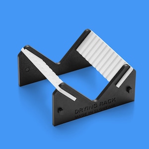 Può includere: Uno stendino in plastica nera con profilo a V e inserti bianchi a coste sui bordi interni. Il lato del supporto presenta il testo in rilievo DRYING RACK e MADE IN SLOVENIA. La struttura è aperta e progettata per sostenere vari oggetti.